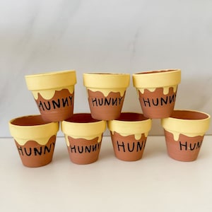 May include: Seven small terracotta pots with yellow paint and the word "Hunny" written in black. The pots are arranged in a semi-circle.