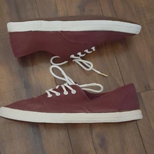 May include: Pair of burgundy canvas sneakers with white rubber soles and white laces. The casual shoes are shown on a wooden surface. The sneakers have a classic, low-profile design.