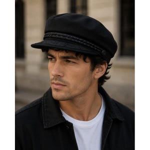 May include: A black fiddler cap featuring a structured crown and a short, stiff brim. The hat is adorned with a decorative braided cord trim that runs across the front, secured on each side by small, circular black buttons with a subtle emblem.