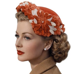 May include: A pillbox hat featuring a vibrant orange felt base adorned with a dense arrangement of velvet flowers in shades of burnt orange and cream. Each flower center is accented with a small, clear, sparkling rhinestone detail.