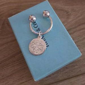 May include: A silver key ring with a circular pendant featuring an intricate design. The key ring is resting on a light blue box with the text "& Co." visible. The key ring has two spherical ends.