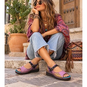 May include: A pair of purple and gold leather sandals featuring a peep-toe design, a black adjustable ankle strap with a metal buckle, and a thick dark sole. The sandals have a multi-toned purple upper with a textured gold panel on the side.