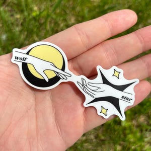 Op de afbeelding: Twee zwart-witte stickers met een hand die uit elk uitsteekt. De sticker links heeft een gele halve maan met het woord "wolf" erop. De sticker rechts heeft een zwarte ster met het woord "star" erop.