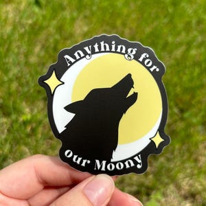May include: A black and white sticker featuring a wolf howling at a yellow crescent moon. The text "Anything for our Moony" is written around the moon.