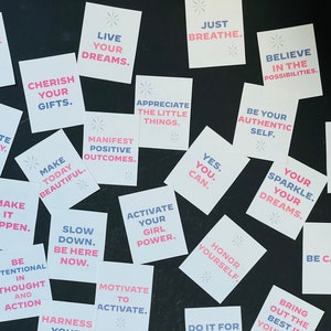 May include: A collection of motivational quotes printed on white cards with pink and blue text. The cards are arranged on a black surface. Some of the quotes include "Live Your Dreams", "Just Breathe", "Believe in the Possibilities", "Make Today Beautiful", "Be Intentional in Thought and Action", "Activate Your Girl Power", "Your Sparkle. Your Dreams.", "Be Calm", and "Bring Out the Best in You".