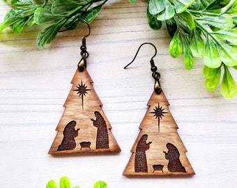 Op de afbeelding: Een paar houten kerstboom oorbellen met een kerststal ontwerp. De oorbellen zijn bruin en hebben een ster bovenop de boom. De kerststal bevat Maria, Jozef en het kindje Jezus in een kribbe.