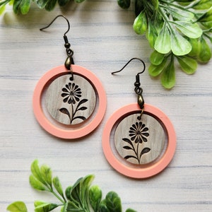 May include: A pair of handmade earrings featuring a floral design. Each earring has a light pink outer circle and a wooden inner circle with a carved flower. The earrings hang from bronze-colored ear wires.