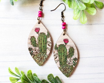 Könnte beinhalten: Ein Paar Holzohrringe mit einem rosa Perlenanhänger. Jeder Ohrring zeigt ein Laser-Cut-Design eines Kaktus und Sukkulenten.
