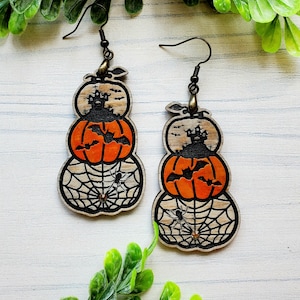 May include: A pair of Halloween-themed earrings featuring stacked wooden pumpkins with a haunted house, bats, and spiderwebs. The pumpkins are painted orange and the spiderwebs are black.