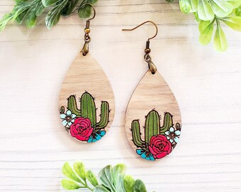 Könnte beinhalten: Ein Paar hölzerne tropfenförmige Ohrringe mit einem bemalten Design aus einem Kaktus und Blumen. Der Kaktus ist grün, die Blumen sind rosa, blau und weiß, und die Ohrringe haben goldene Haken.