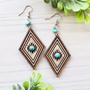 May include: A pair of wooden earrings with a turquoise stone in the center of each earring. The earrings are shaped like a diamond and have a geometric pattern carved into the wood.