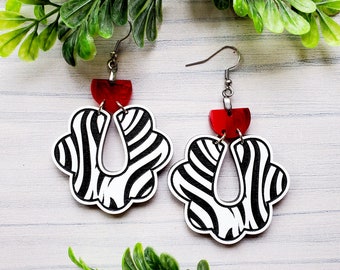 May include: A pair of black and white zebra print earrings with red accents. The earrings are made of wood and have a unique shape.