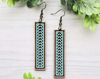 May include: A pair of wooden earrings with a geometric pattern. The earrings are painted a light blue color and have a brown wood frame. The earrings are hanging from gold-colored hooks.