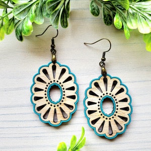 Puede incluir: Un par de pendientes de madera con un borde azul turquesa. Los pendientes tienen forma de flor con un recorte circular en el centro.