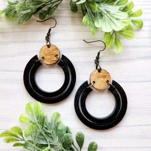 May include: A pair of black wooden hoop earrings with a natural wood circle accent. The earrings have a simple, modern design.
