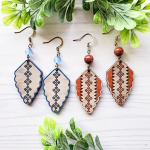 May include: Three pairs of wooden earrings with geometric patterns. The earrings are in blue, orange, and brown. Each pair has a different colored bead.