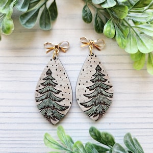 May include: A pair of wooden dangle earrings with a green pine tree design on a white background. The earrings have gold bows at the top.
