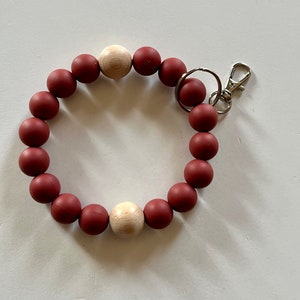 May include: A burgundy beaded keychain bracelet with two natural wood beads. The bracelet has a silver-colored clasp and key ring. The beads are arranged in a circular pattern.