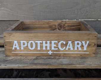 May include: A rustic wooden box with the word "APOTHECARY" painted in white on the front. The box has a natural wood finish and is perfect for storing small items.