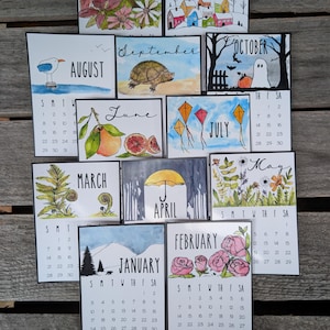 Puede incluir: Un conjunto de doce tarjetas de calendario mensual individual con ilustraciones en acuarela para cada mes, incluyendo enero con montañas, febrero con rosas, marzo con helechos, abril con un paraguas, mayo con flores silvestres, junio con cítricos, julio con cometas, agosto con una gaviota, septiembre con una tortuga, octubre con un fantasma, noviembre con flores y diciembre con casas.