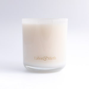 May include: A clear glass candle jar with a white candle inside. The candle has a gold label that reads "Labor & Mirth".