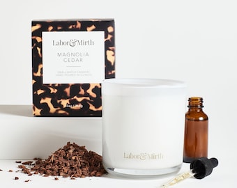 May include: A white candle with a clear glass jar and a black and brown patterned box. The candle is labeled "Labor & Mirth Magnolia Cedar" and "13oz/368g". The candle is next to a small brown glass bottle with a dropper.