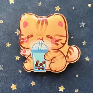May include: A cute orange tabby cat with pink cheeks is holding a blue and white boba tea cup with brown boba pearls. The cat is smiling and has a straw in its mouth.