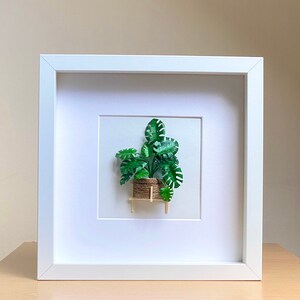 May include: A white framed picture with a green paper plant in a brown pot on a small wooden stand. The plant is a monstera deliciosa.