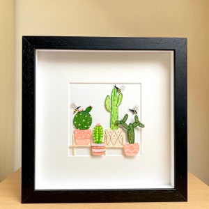 May include: A framed artwork featuring a paper craft scene of five potted cacti with bees flying around them. The cacti are in different sizes and shapes, and the pots are pink and white. The background is white, and the frame is black.