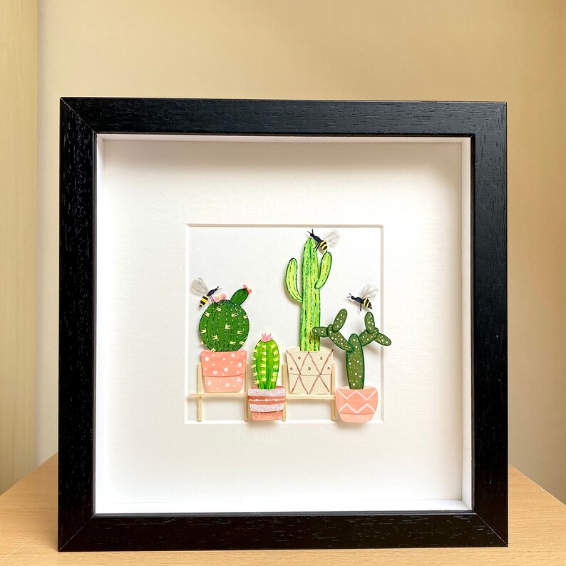 May include: A framed artwork featuring a paper craft scene of five potted cacti with bees flying around them. The cacti are in different sizes and shapes, and the pots are pink and white. The background is white, and the frame is black.