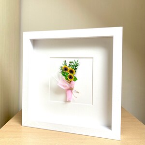 May include: A white frame with a white mat and a small bouquet of yellow sunflowers with green leaves tied with a pink ribbon.