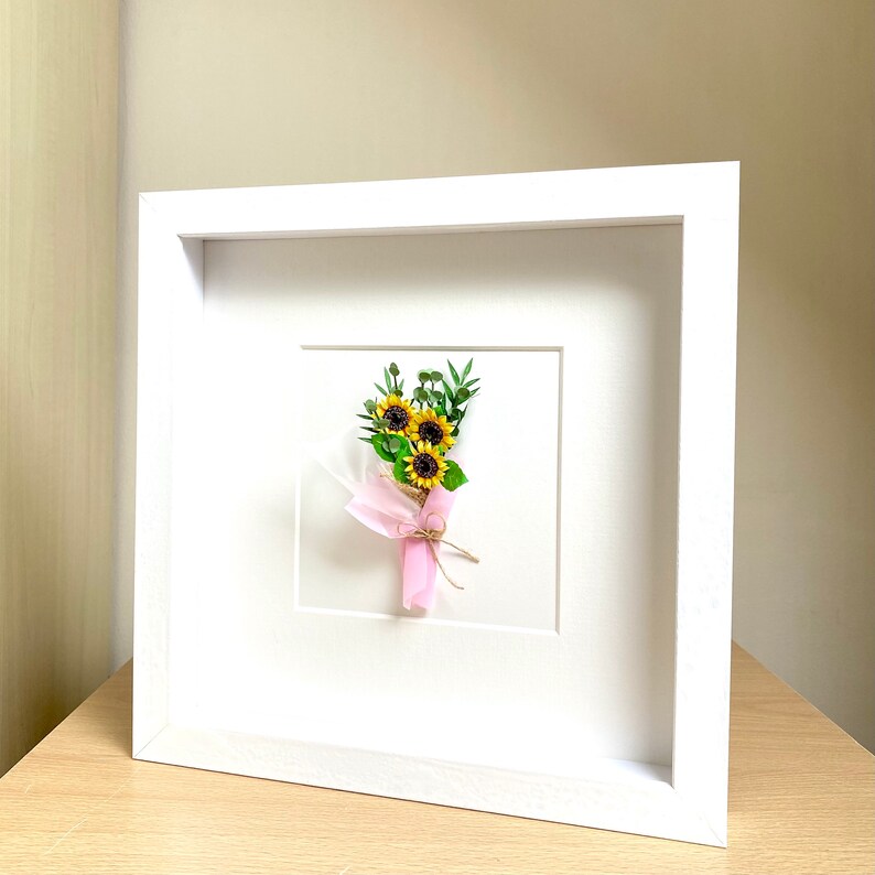 May include: A white frame with a white mat and a small bouquet of yellow sunflowers with green leaves tied with a pink ribbon.