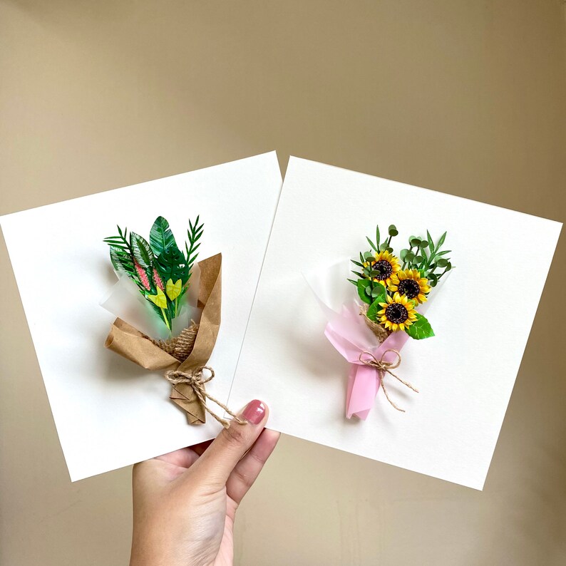 May include: Two handmade paper flower bouquets on white cards. One bouquet is made of green leaves and yellow flowers, the other is made of yellow sunflowers and green leaves. Both bouquets are wrapped in brown paper and tied with twine.
