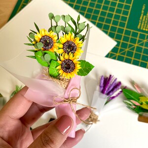 May include: A small bouquet of paper sunflowers wrapped in pink tissue paper and tied with twine. The bouquet is held in a hand.