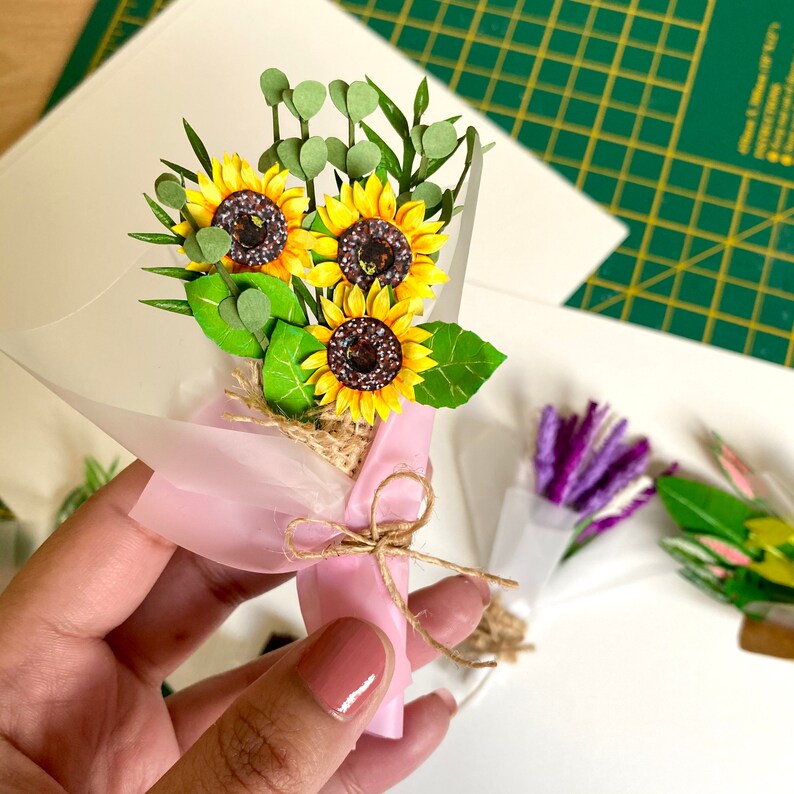 May include: A small bouquet of paper sunflowers wrapped in pink tissue paper and tied with twine. The bouquet is held in a hand.