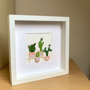 May include: A white framed picture with a white mount and a beige background. The picture features five small potted cacti with pink pots and green cacti. The cacti are labelled with the names Wendy, Liam, Ness, and Joe. There are small yellow and black bees flying around the cacti.