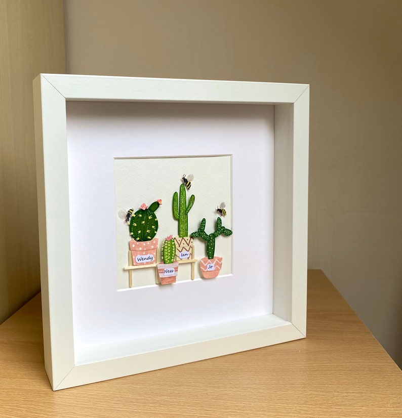 May include: A white framed picture with a white mount and a beige background. The picture features five small potted cacti with pink pots and green cacti. The cacti are labelled with the names Wendy, Liam, Ness, and Joe. There are small yellow and black bees flying around the cacti.