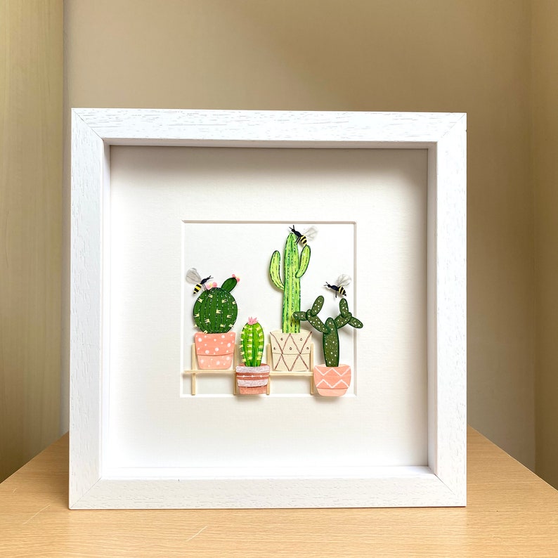 May include: A white framed picture with a white mount and a white background. The picture features a collection of five potted cacti with pink pots and green cacti. There are three yellow and black bees flying around the cacti.