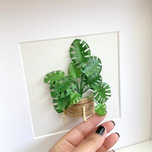 May include: A framed papercraft artwork featuring a green monstera plant in a brown woven basket with wooden legs. The plant is made of paper and is displayed on a white background.