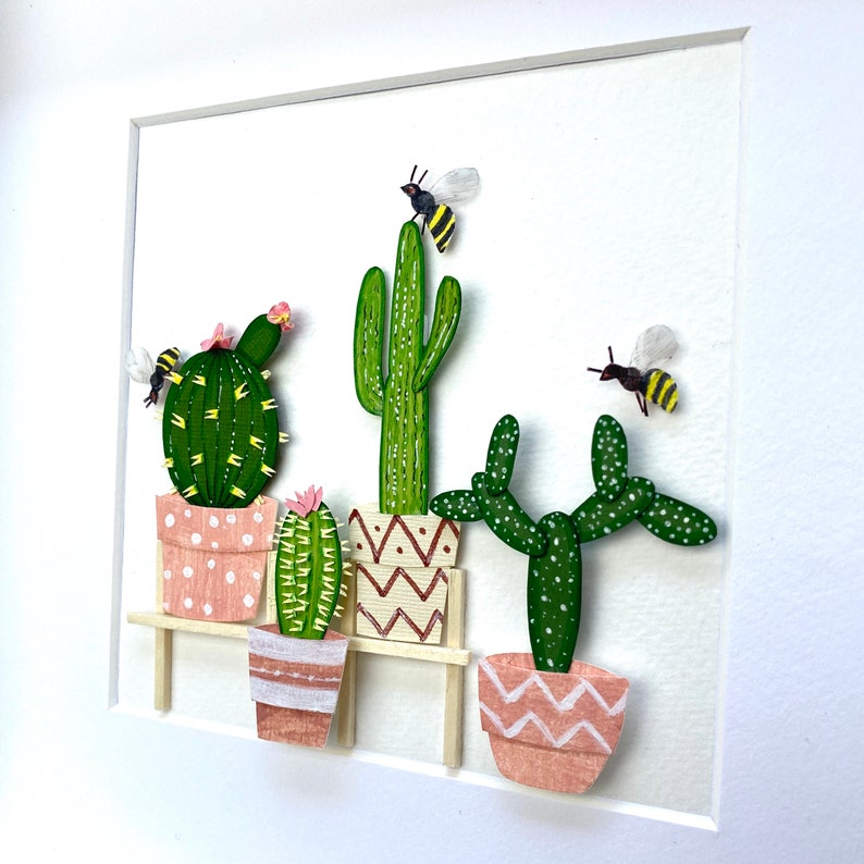 May include: A framed artwork featuring a collection of potted cacti and succulents with a wooden shelf. The cacti are green with pink and white pots. There are three bees flying around the cacti.