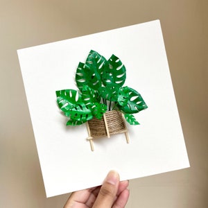 May include: A papercraft of a potted plant with green leaves and a brown rope-wrapped pot on a wooden stand. The plant is on a white background.