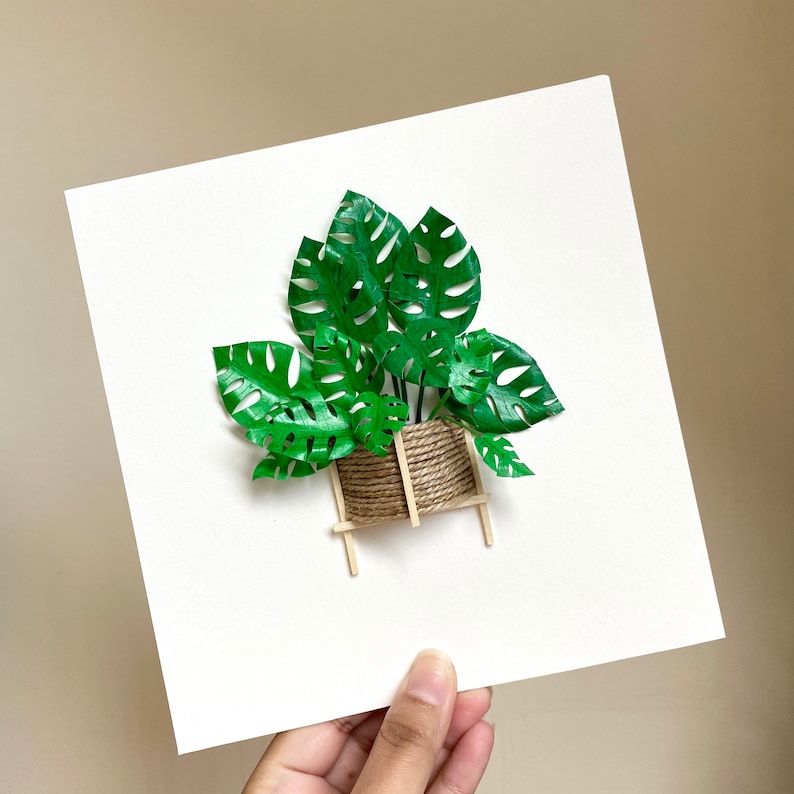 May include: A papercraft of a potted plant with green leaves and a brown rope-wrapped pot on a wooden stand. The plant is on a white background.
