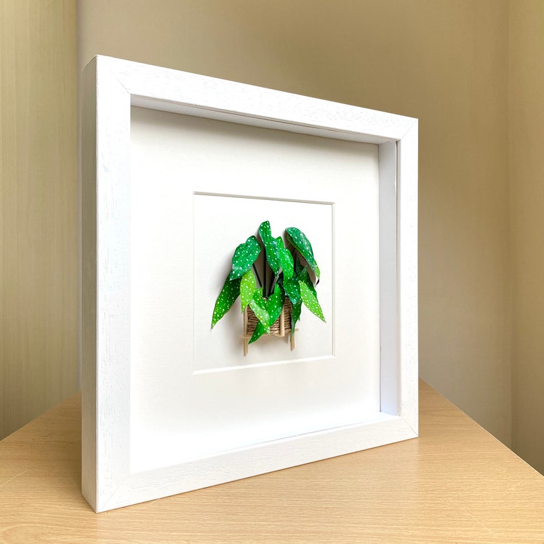 May include: White frame with a green paper plant inside. The plant is in a small brown basket and has white dots on the leaves.