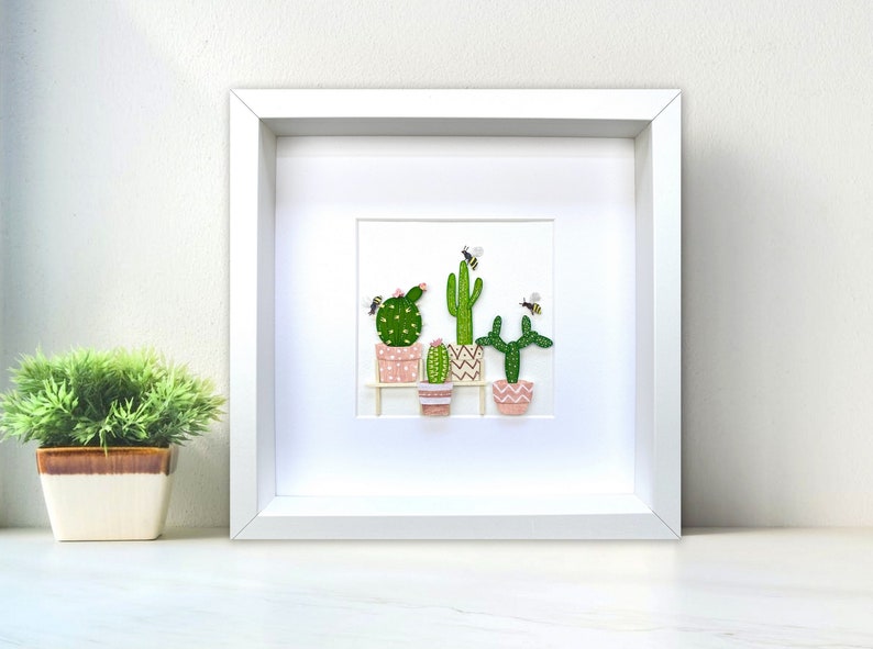 May include: A framed artwork featuring a collection of potted cacti and succulents with pink and white patterned pots.  The cacti are green and have small white flowers.  There are small yellow bees flying around the cacti.