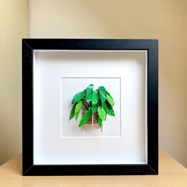 May include: A framed papercraft artwork of a green plant with white dots, in a brown pot, against a white background.