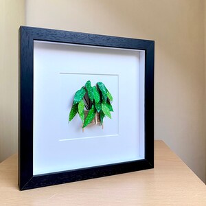 May include: A framed print of a green and white spotted plant in a woven basket. The print is in a black frame with a white mat.