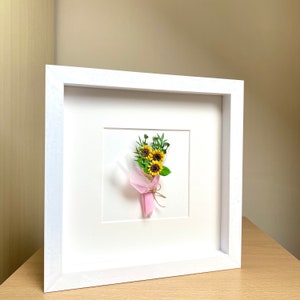May include: A white frame with a small bouquet of paper sunflowers tied with twine and a pink tissue paper wrapper.