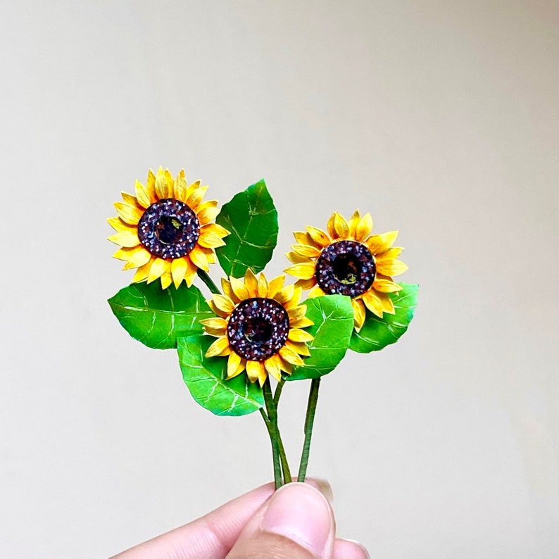 May include: Three paper sunflowers with yellow petals and brown centers are arranged on green stems with leaves. The sunflowers are crafted from paper and have a realistic appearance.