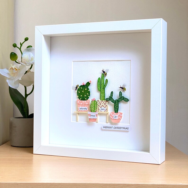 May include: A white framed picture with a white background featuring a collection of five potted cacti with pink and green pots. The cacti are labelled "Wendy", "Ness", "Liam", and "Jo". The picture also includes three bees and the text "Merry Christmas".
