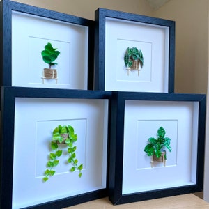 May include: Four black framed pictures with white mats and a variety of green paper plants in each. The plants are in small pots and are on small wooden stands.